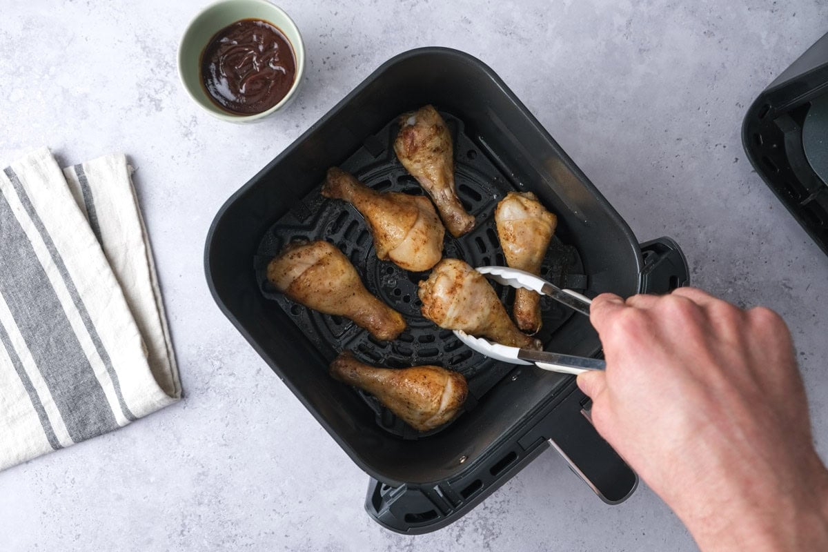 hand with tongs turning raw chicken drumsticks in air fryer basket on counter.