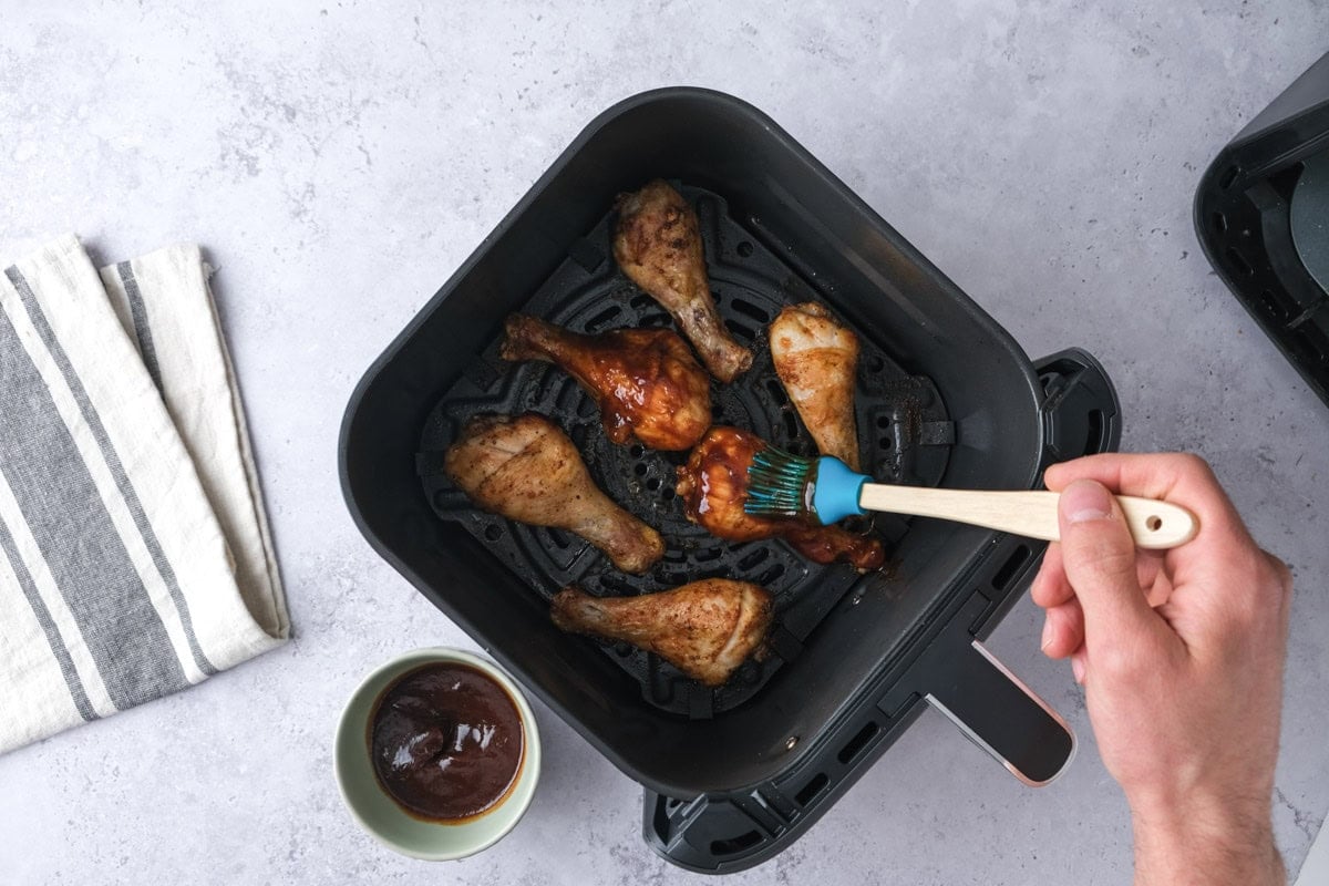 hand brushing bbq sauce onto cooked chicken drumsticks in air fryer basket.