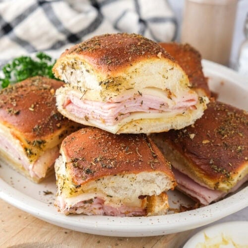 ham and cheese sliders stacked on plate on wooden board with cloth behind.