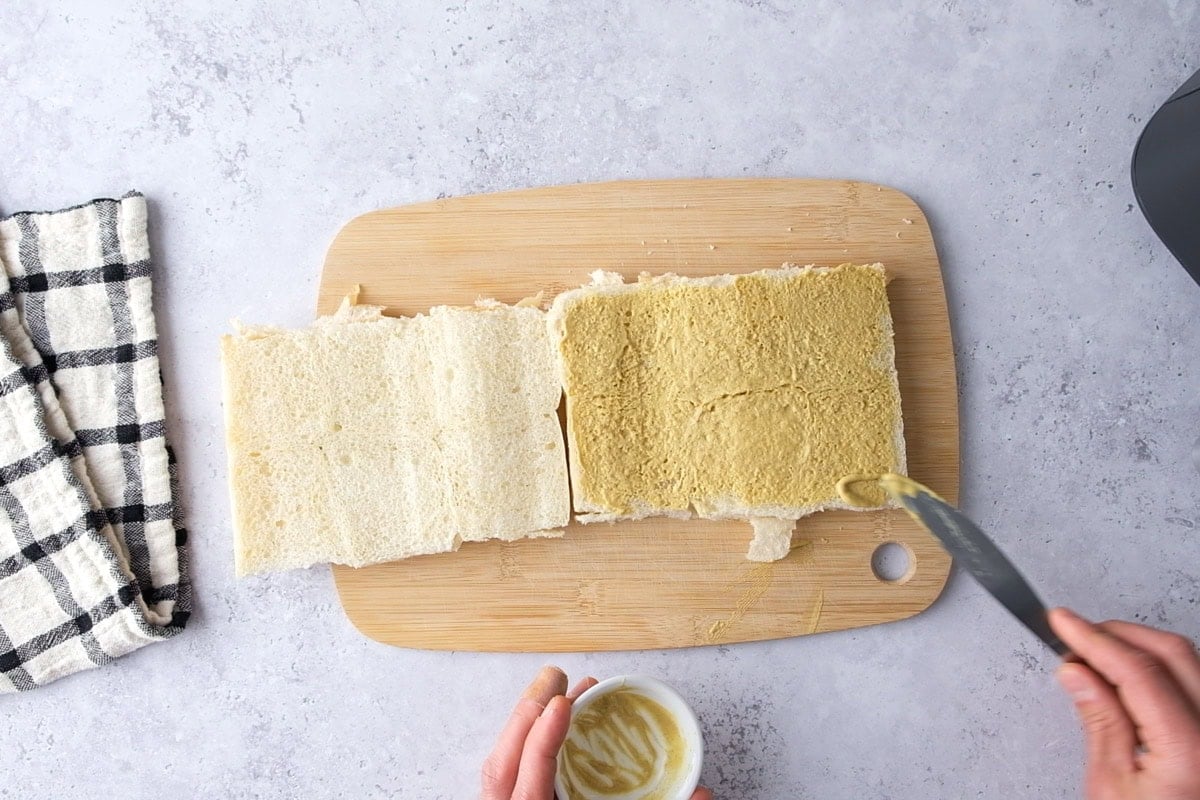 hand with knife spreading mustard on slider buns on cutting board.