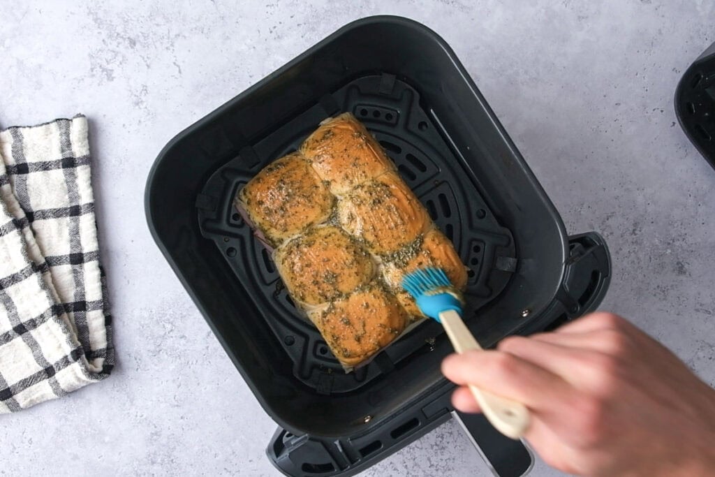 hand using silicone brush to spread butter mixture onto slider buns in air fryer.