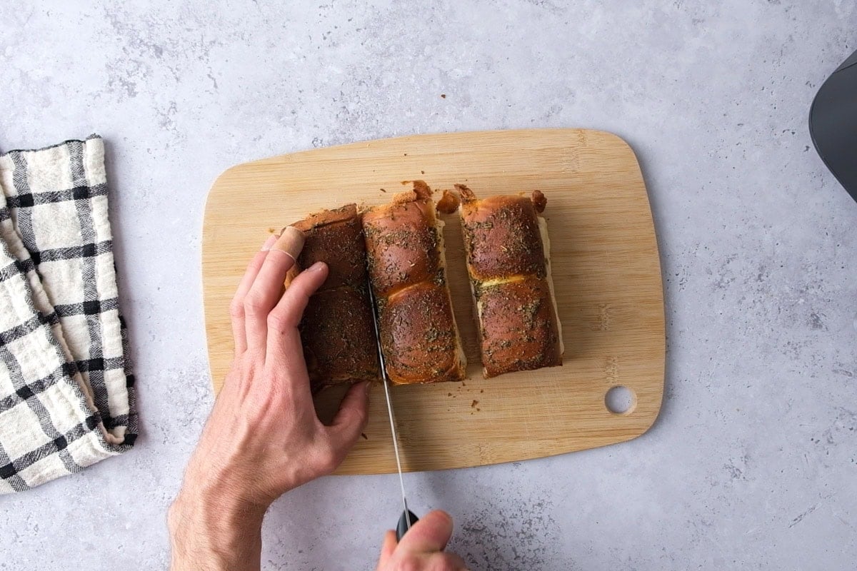 hand cutting crispy ham and cheese sliders with large knife on cutting board.