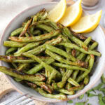 bowl of lemon garlic green beans on wooden board with lemon wedges behind.