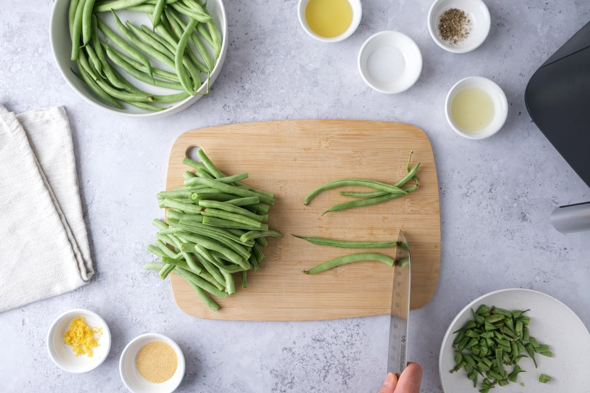 hand holding knife cutting green beans on cutting board with ingredients around.