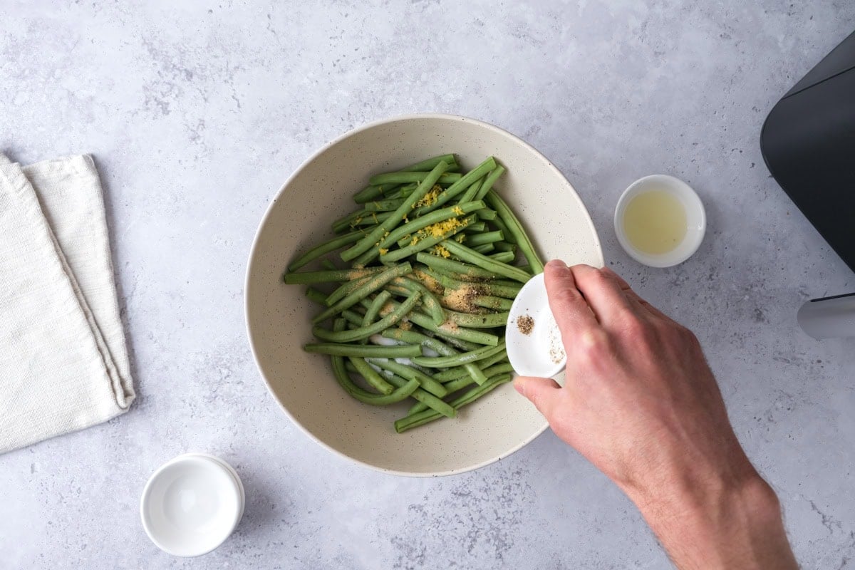 hand pouring ramekin of pepper into bowl of raw green beans and spices on counter.