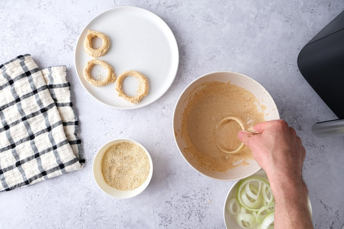 hand dipping onion ring into bowl of batter with breaded rings beside on plate.