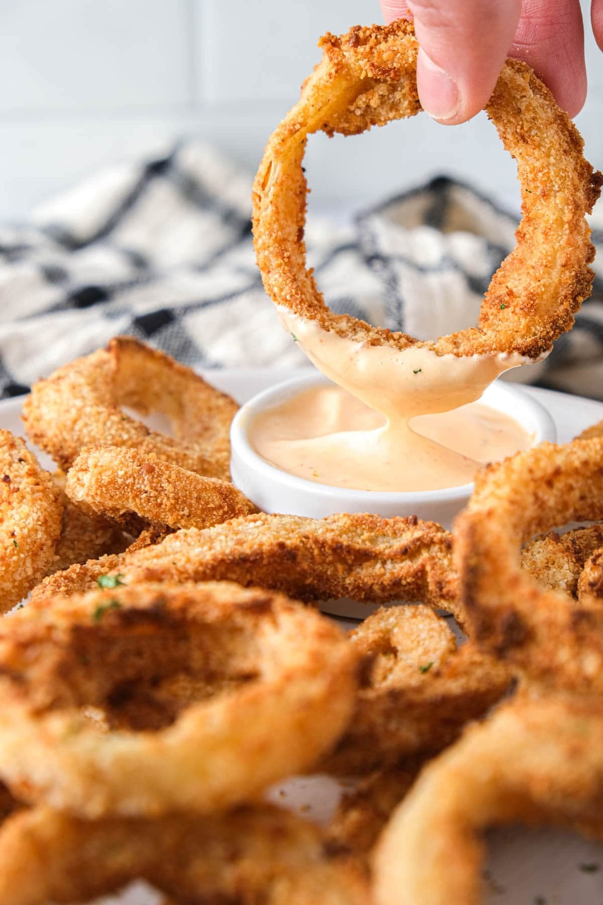 crispy onion ring being dipped into spicy sauce with more on the plate.