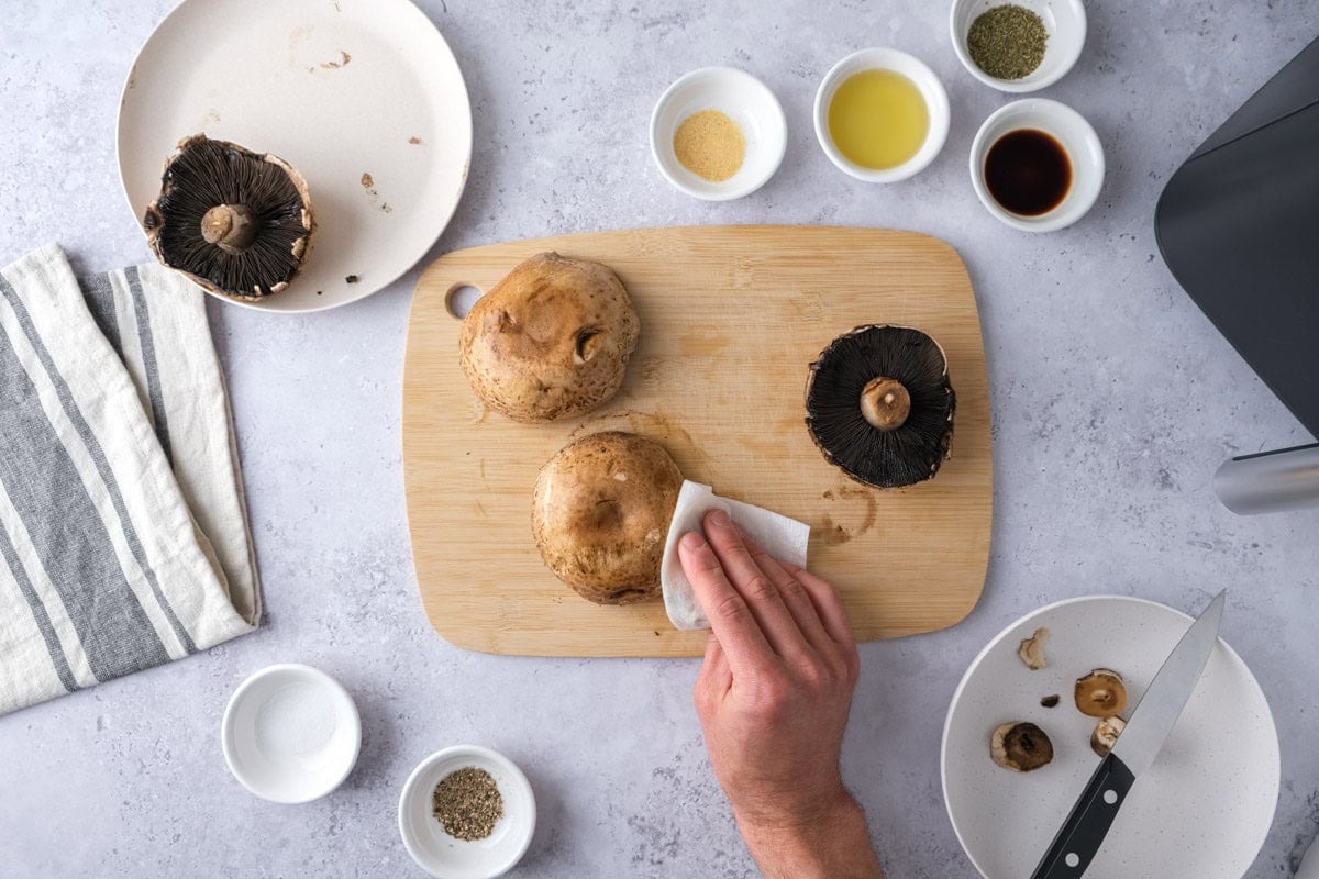 hand cleaning portobello mushrooms with paper towel on counter with ingredients around.