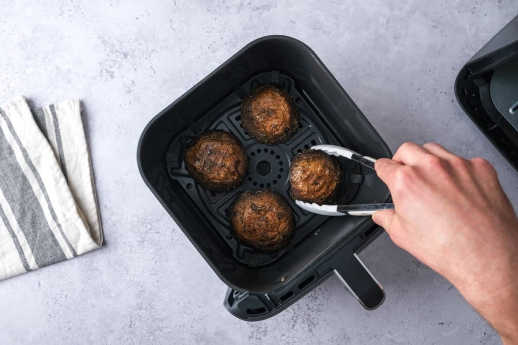 hand with white tongs flipping over portobello mushrooms in air fryer basket.