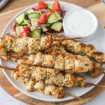 cooked chicken skewers on plate with greek salad and tzatziki on plate behind.