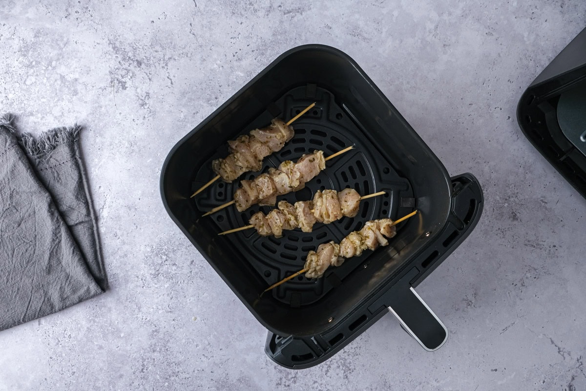 raw chicken souvlaki skewers in air fryer basket on counter.
