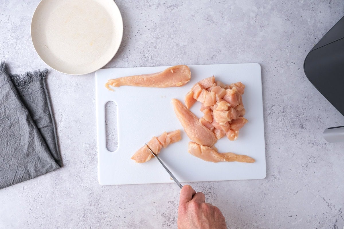 hand cutting pieces of chicken with knife on white cutting board.