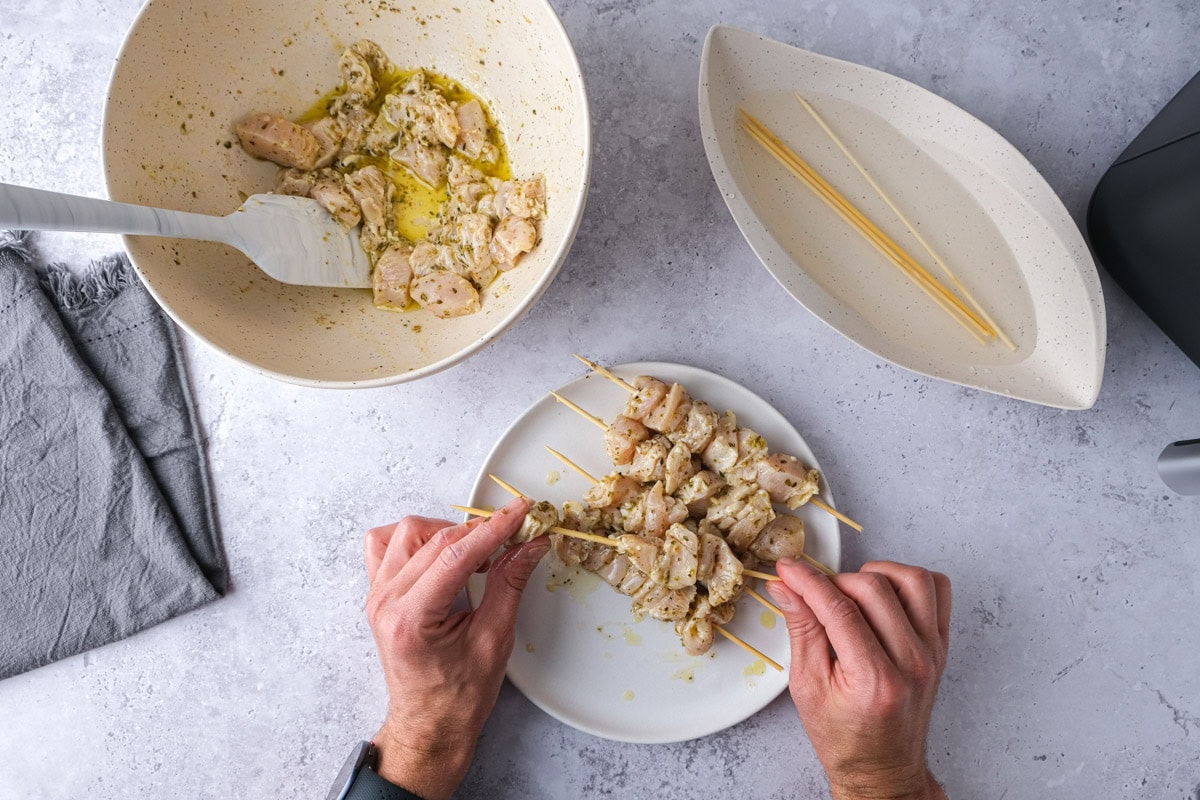 hand placing raw chicken pieces on wooden skewers.