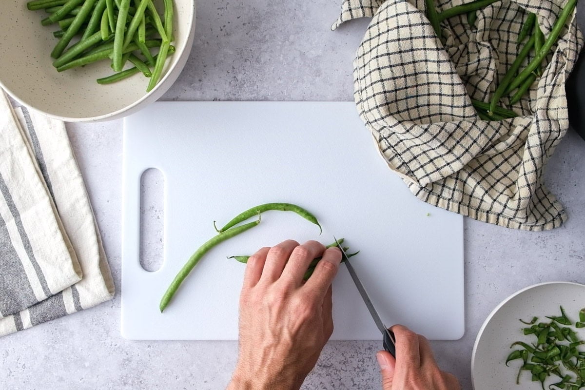 hand cutting green beans with knife on cutting board.