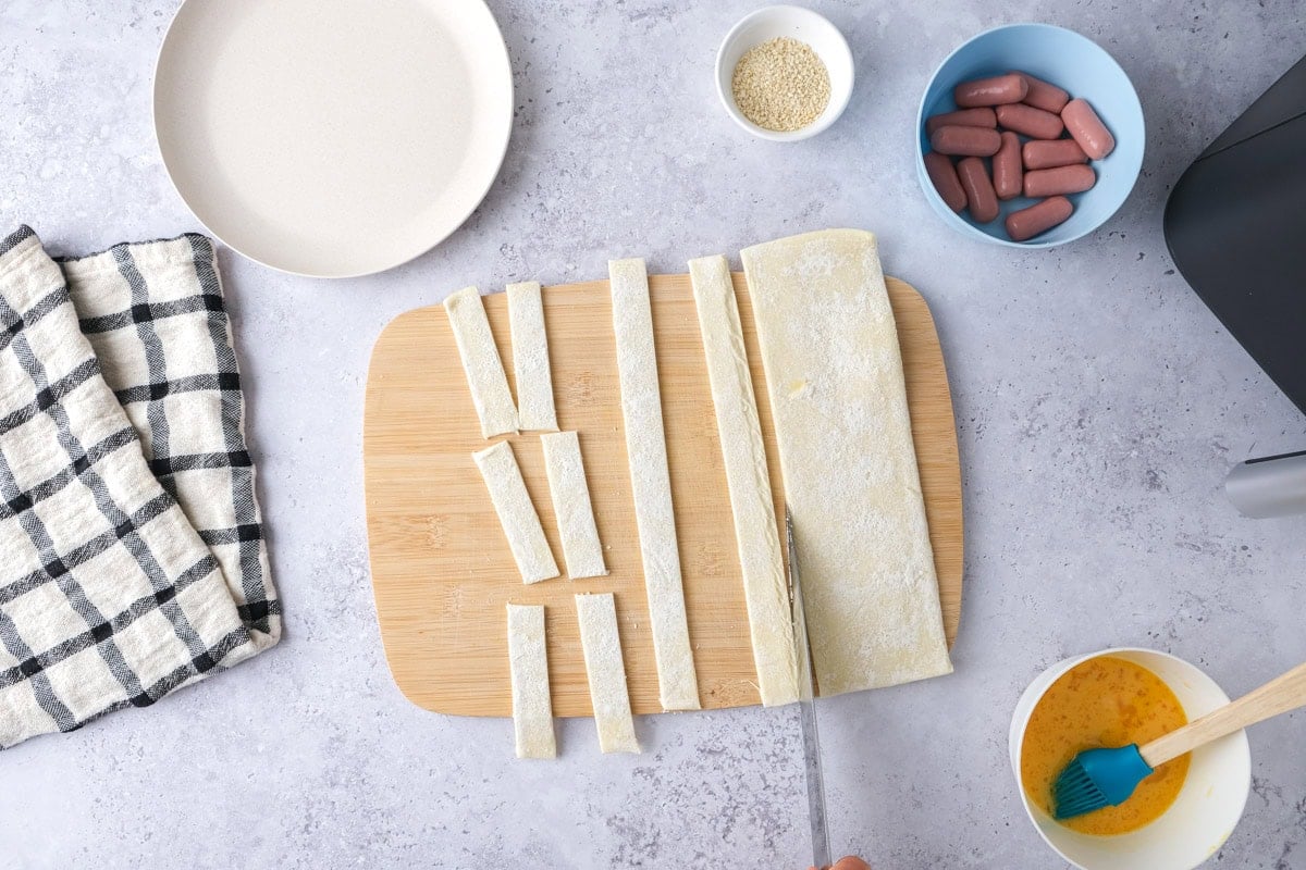 hand with knife cutting puff pastry into strips on cutting board.