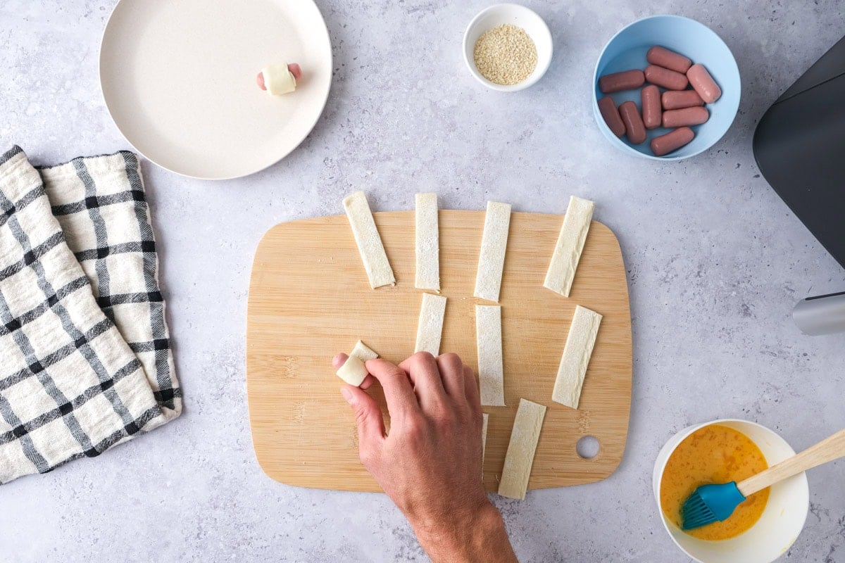 hand rolling mini sausages into puff pastry on cutting board.