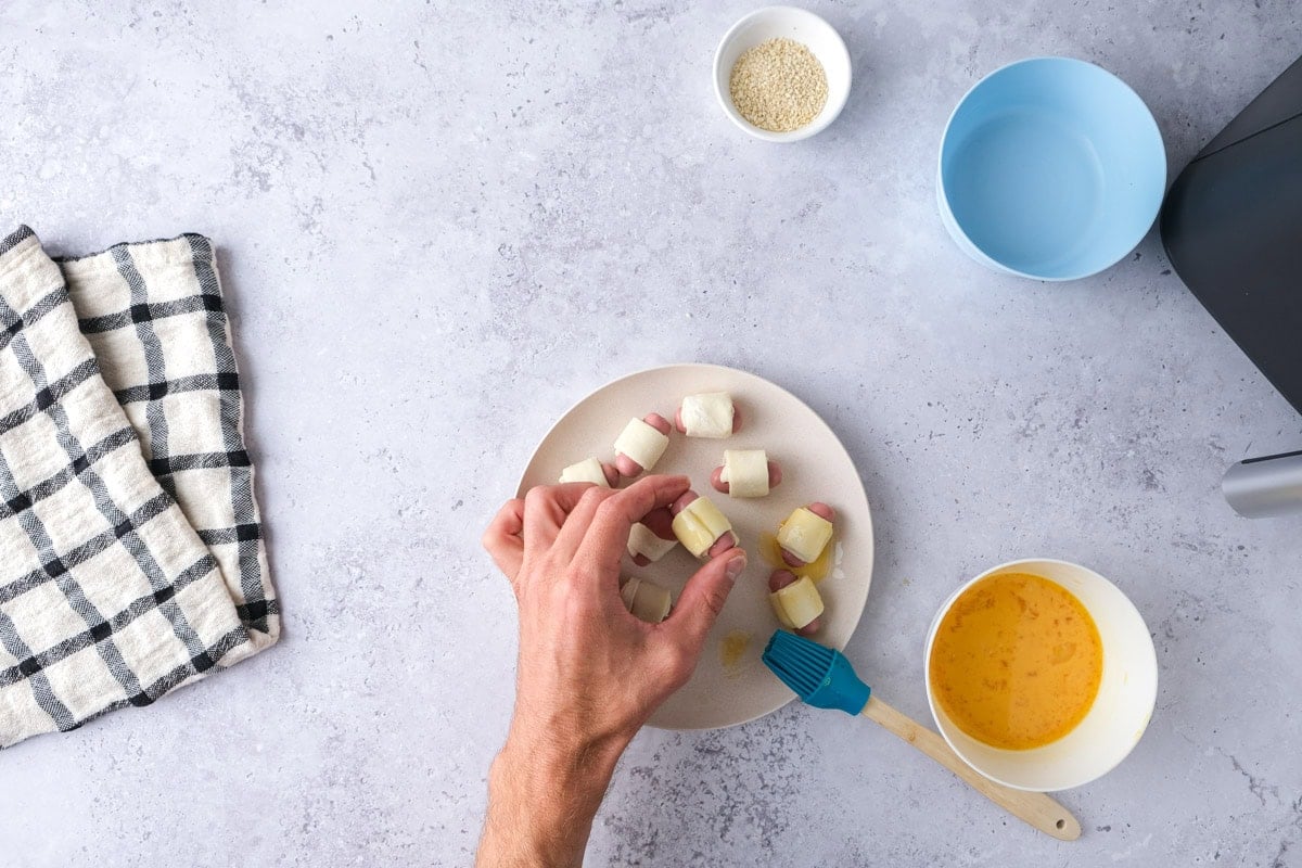 hand adding egg wash to raw pigs in a blanket on plate.