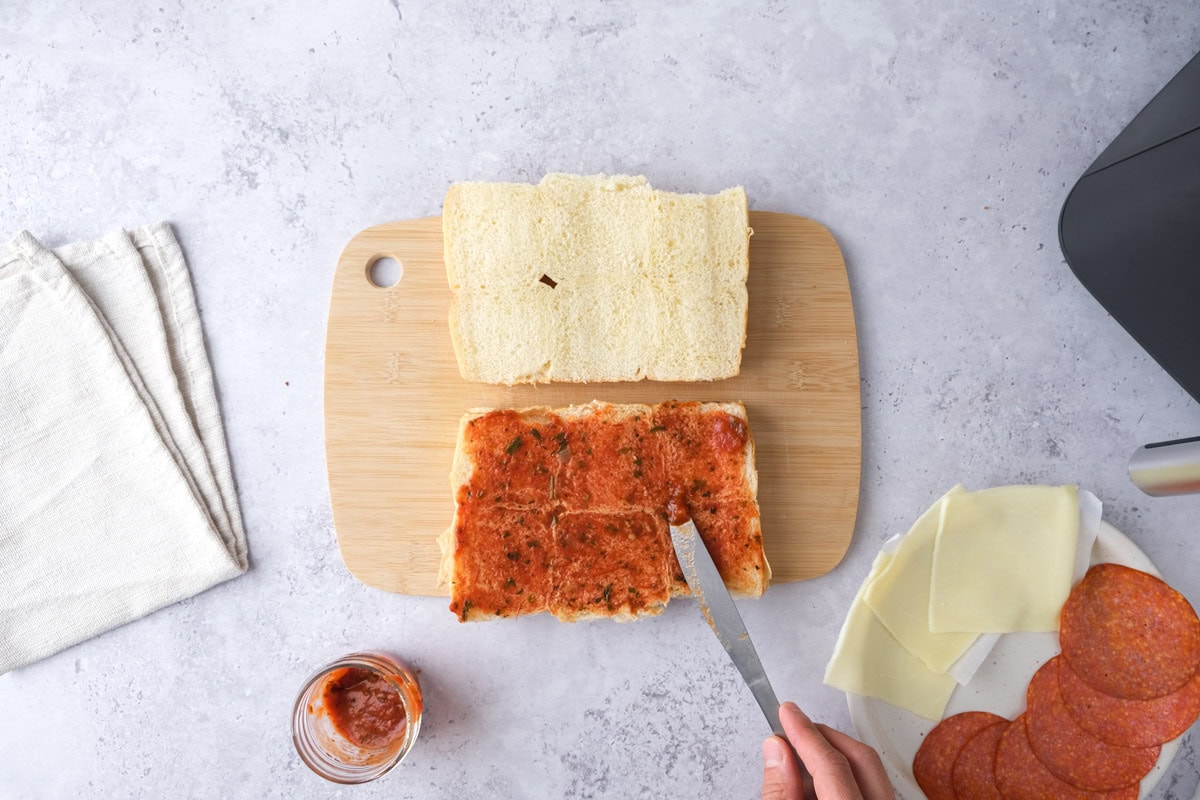 hand with knife spreading pizza sauce on bottom of slider buns.