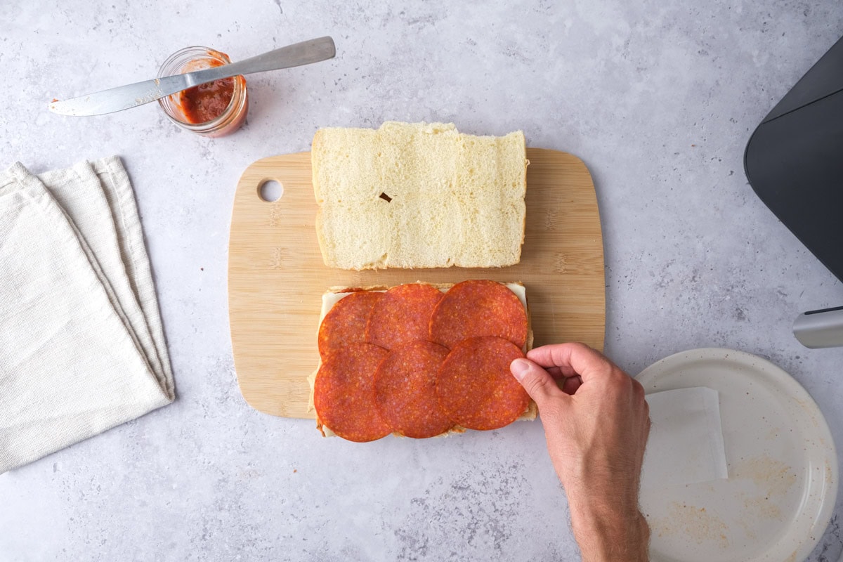 hand placing deli pepperoni on slider buns on wooden cutting board.
