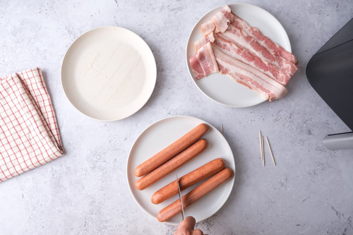 knife scoring hot dogs on a plate with raw bacon beside on counter.