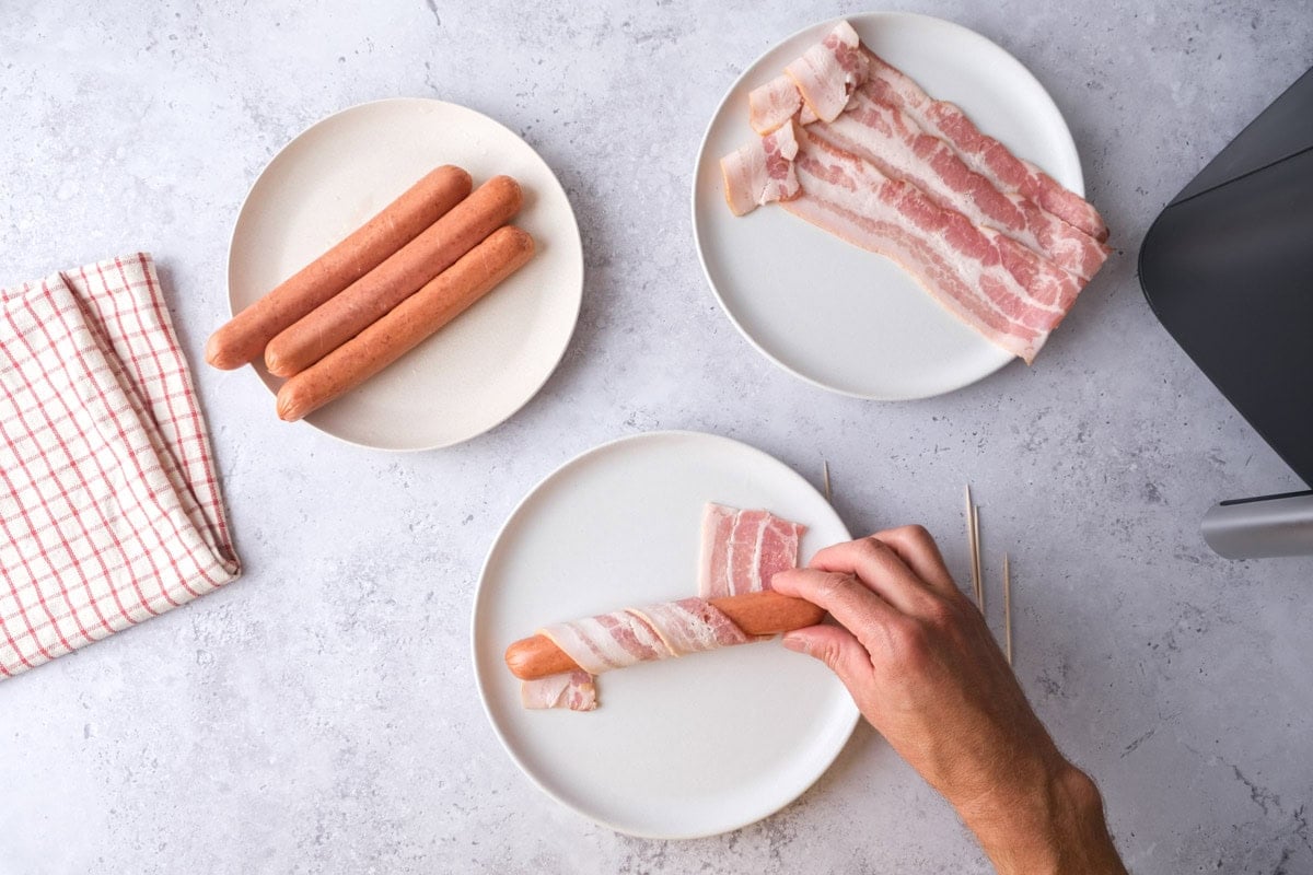 hand wrapping bacon strip around a hot dog on plate on counter.