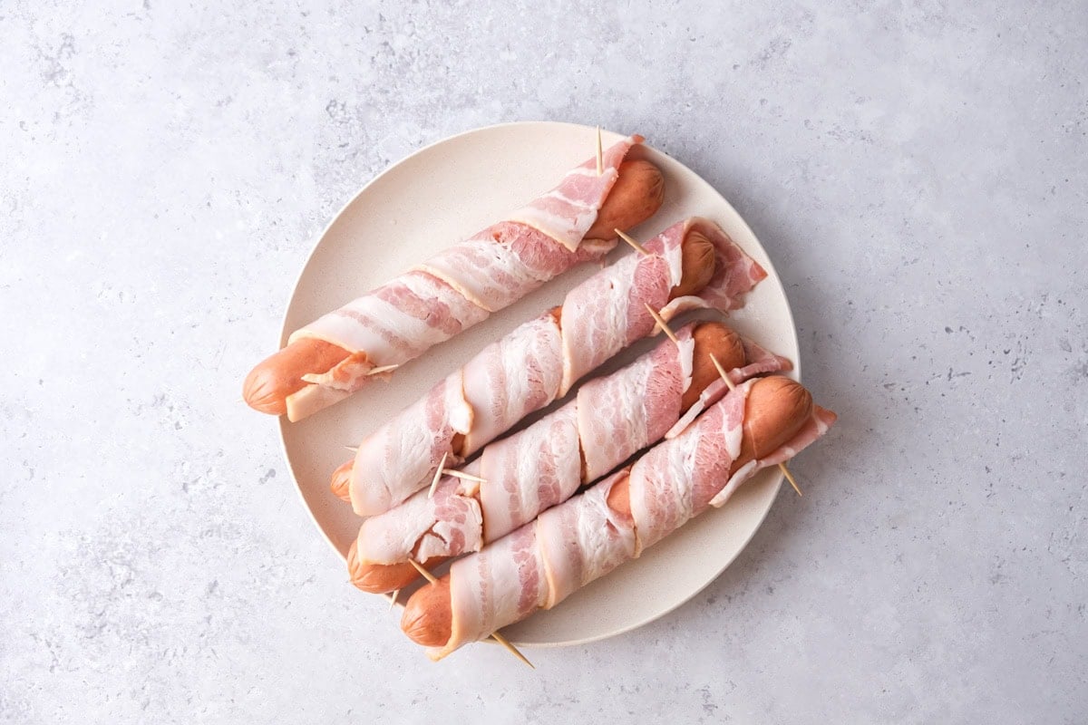four raw bacon wrapped hot dogs on a plate on counter.