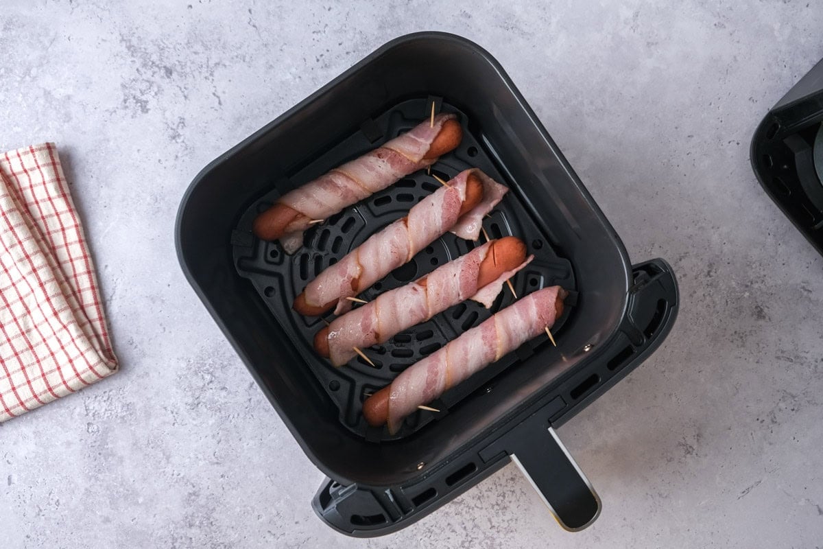 raw bacon wrapped hot dogs in black air fryer basket on counter.