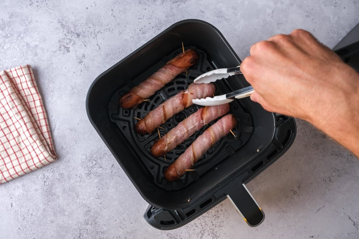 tongs flipping bacon wrapped hot dogs in air fryer basket on counter.
