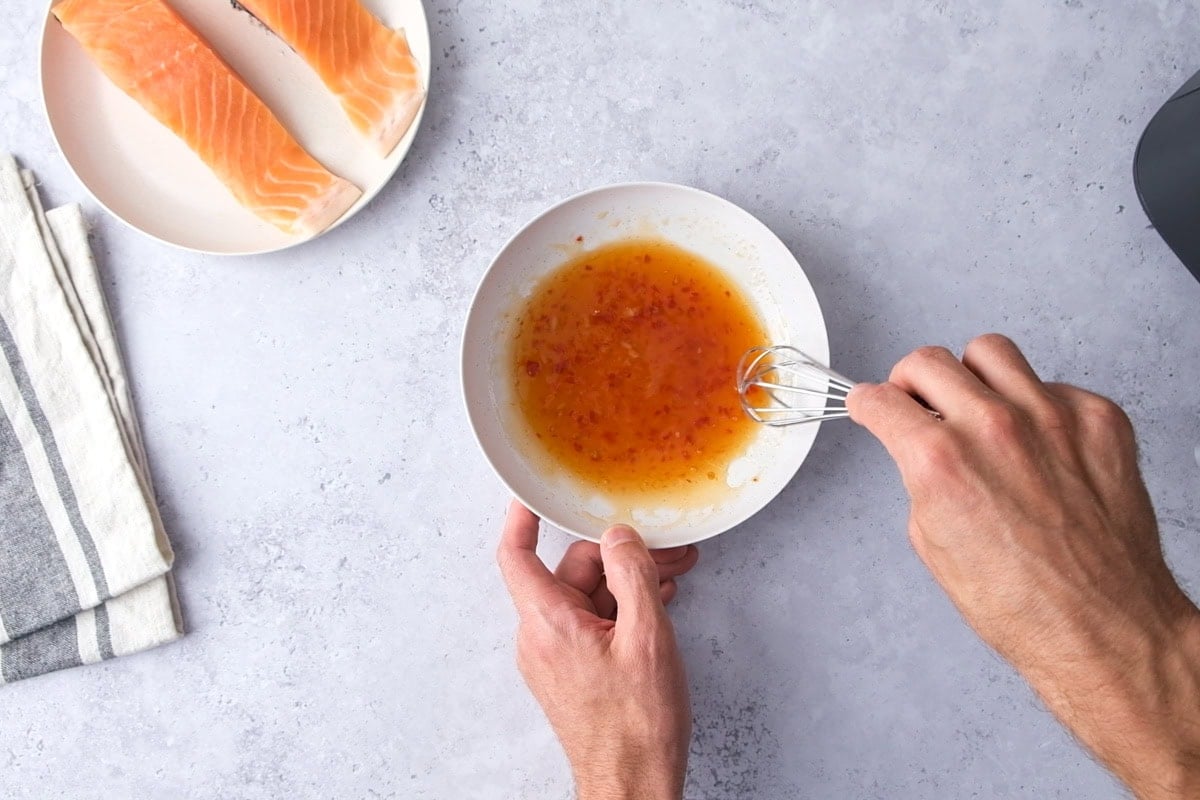 hand mixing sweet chili marinade in bowl with salmon fillets beside.