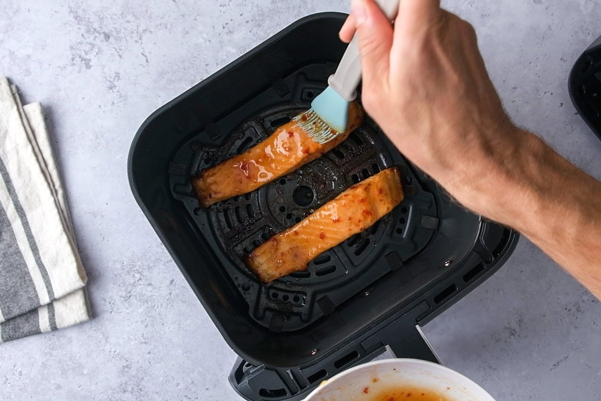 hand with silicone brush brushing marinade onto salmon fillets in air fryer basket.