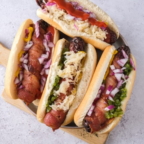 bacon wrapped hot dogs in buns with condiments on plate on counter.
