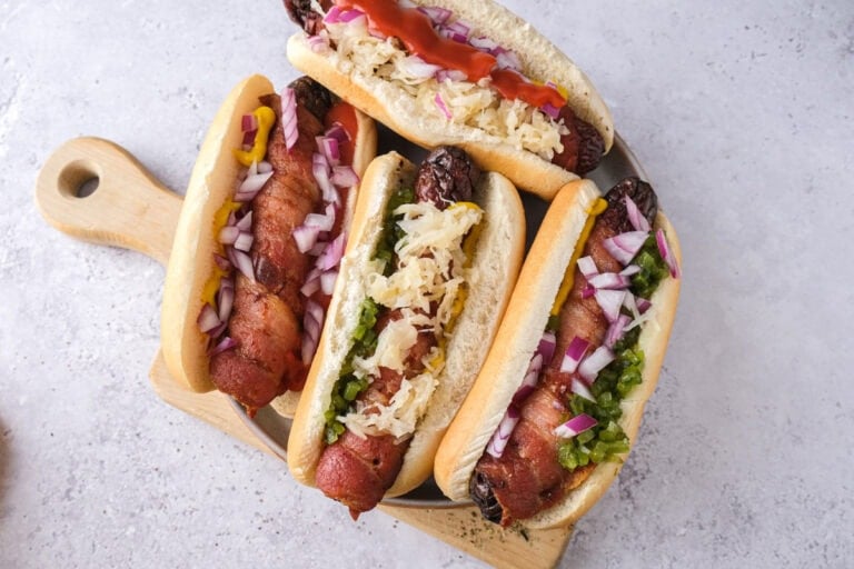 bacon wrapped hot dogs in buns with condiments on plate on counter.