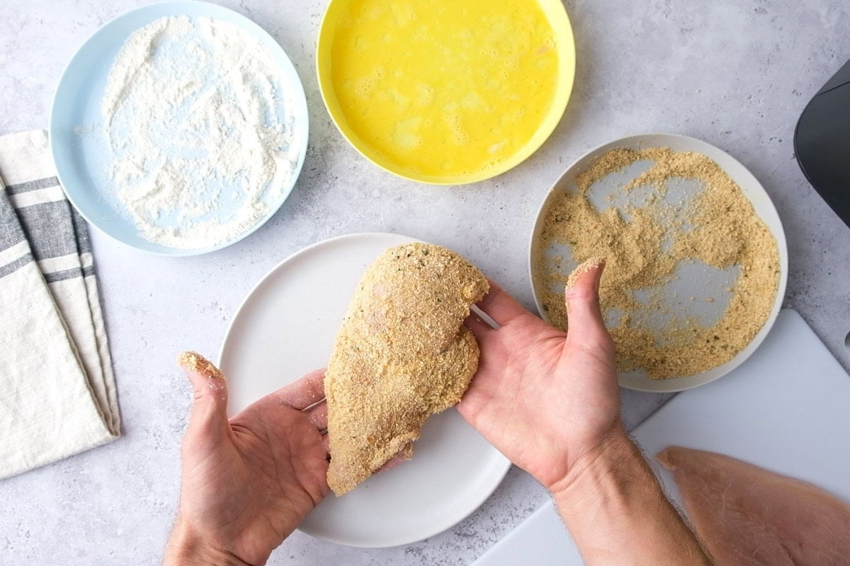hand holding freshly breaded chicken breast with ingredients around.