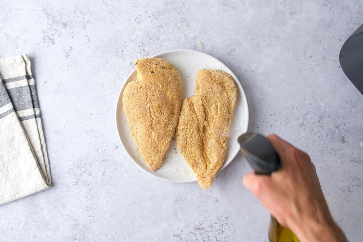 breaded chicken breasts on a plate being sprayed with oil spray.