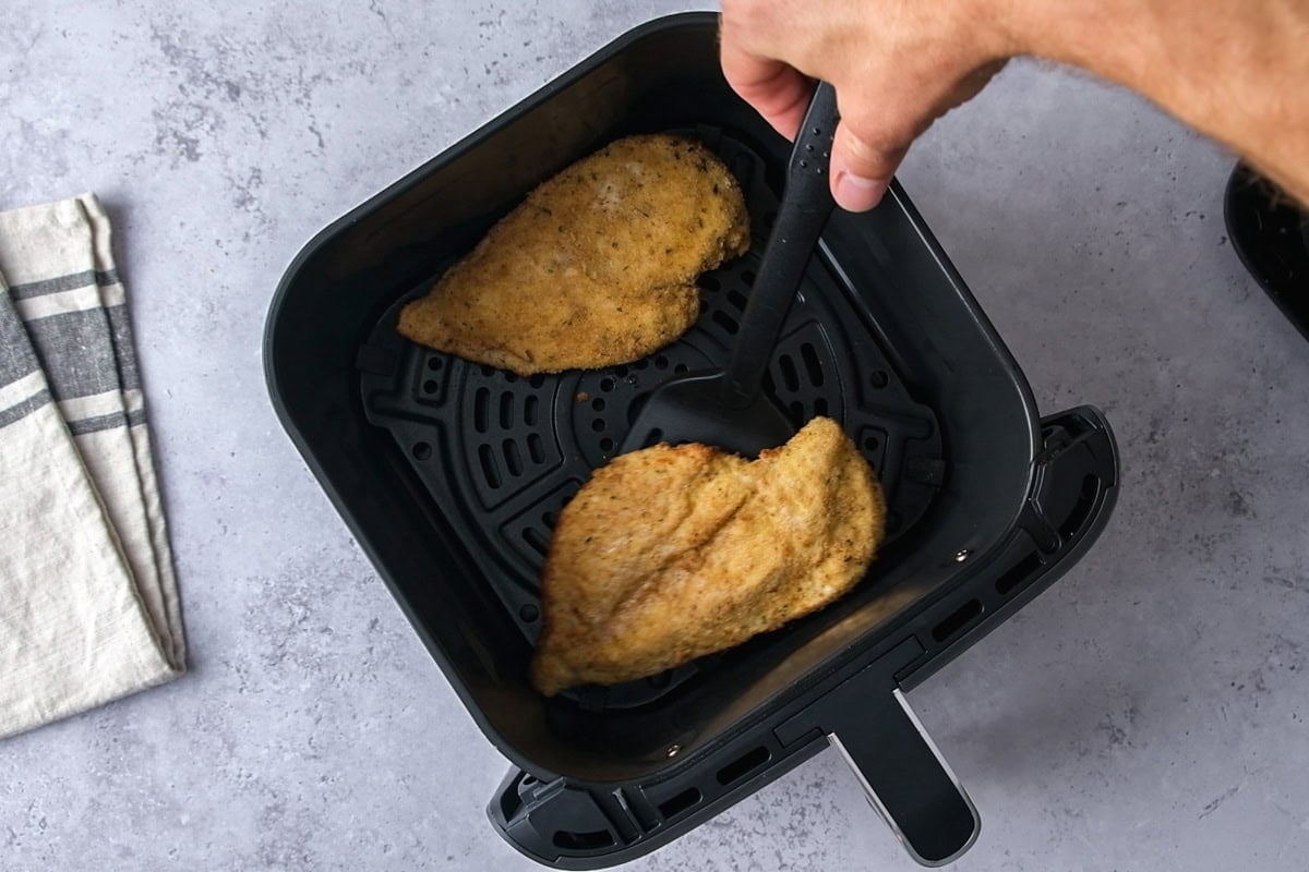 hand with flipper flipping cooked chicken breasts in air fryer basket.