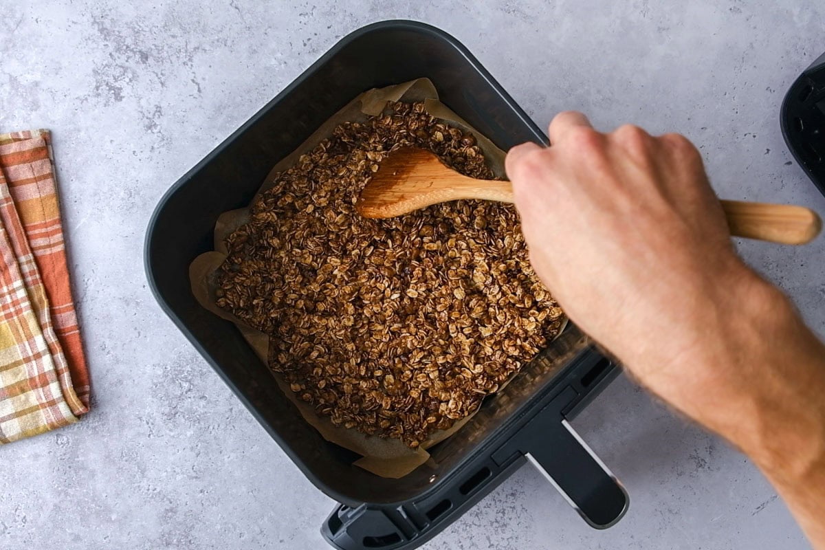 wooden spoon moving granola in bottom of air fryer basket on counter.