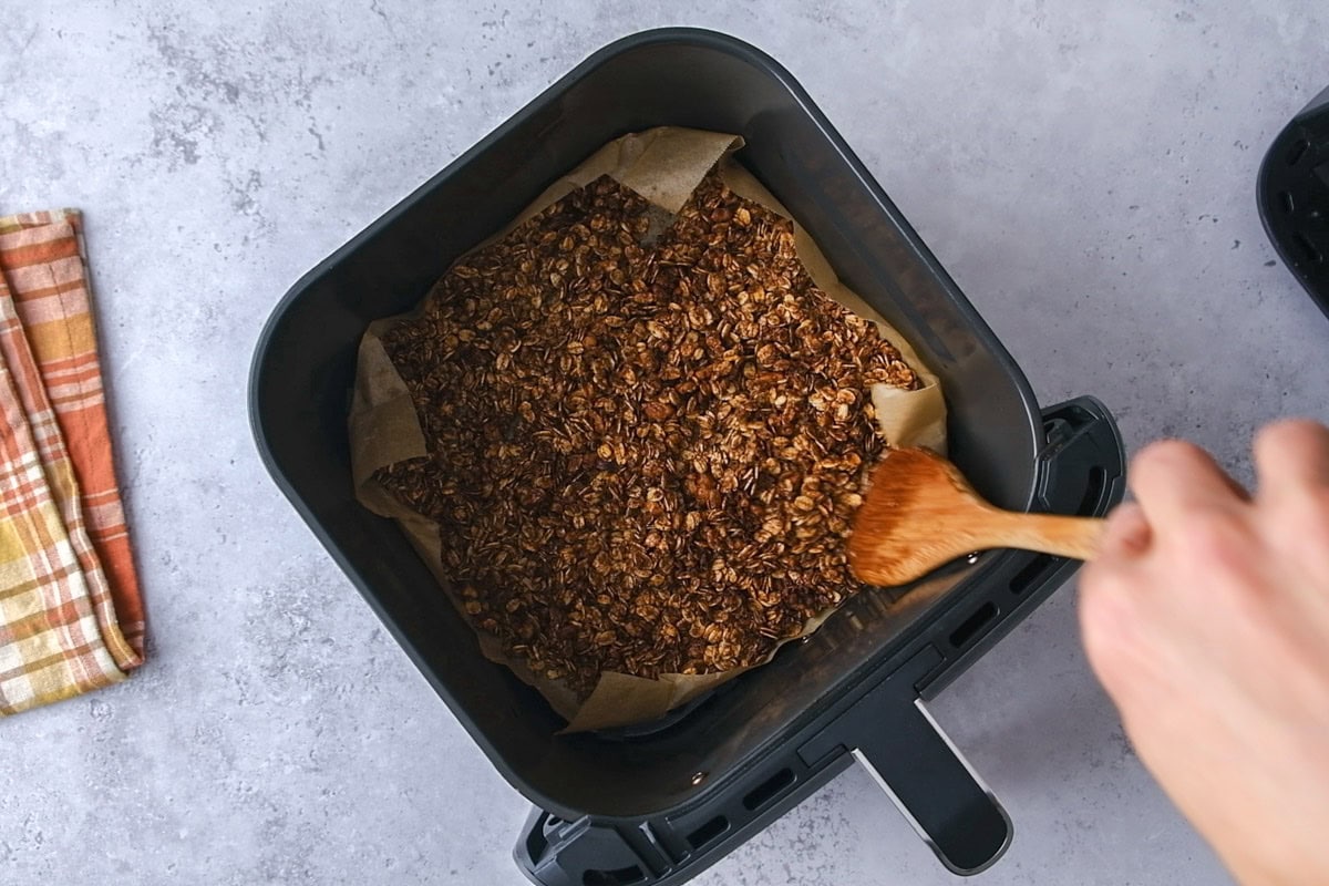 raw granola in bottom of air fryer basket with wooden spoon stirring.